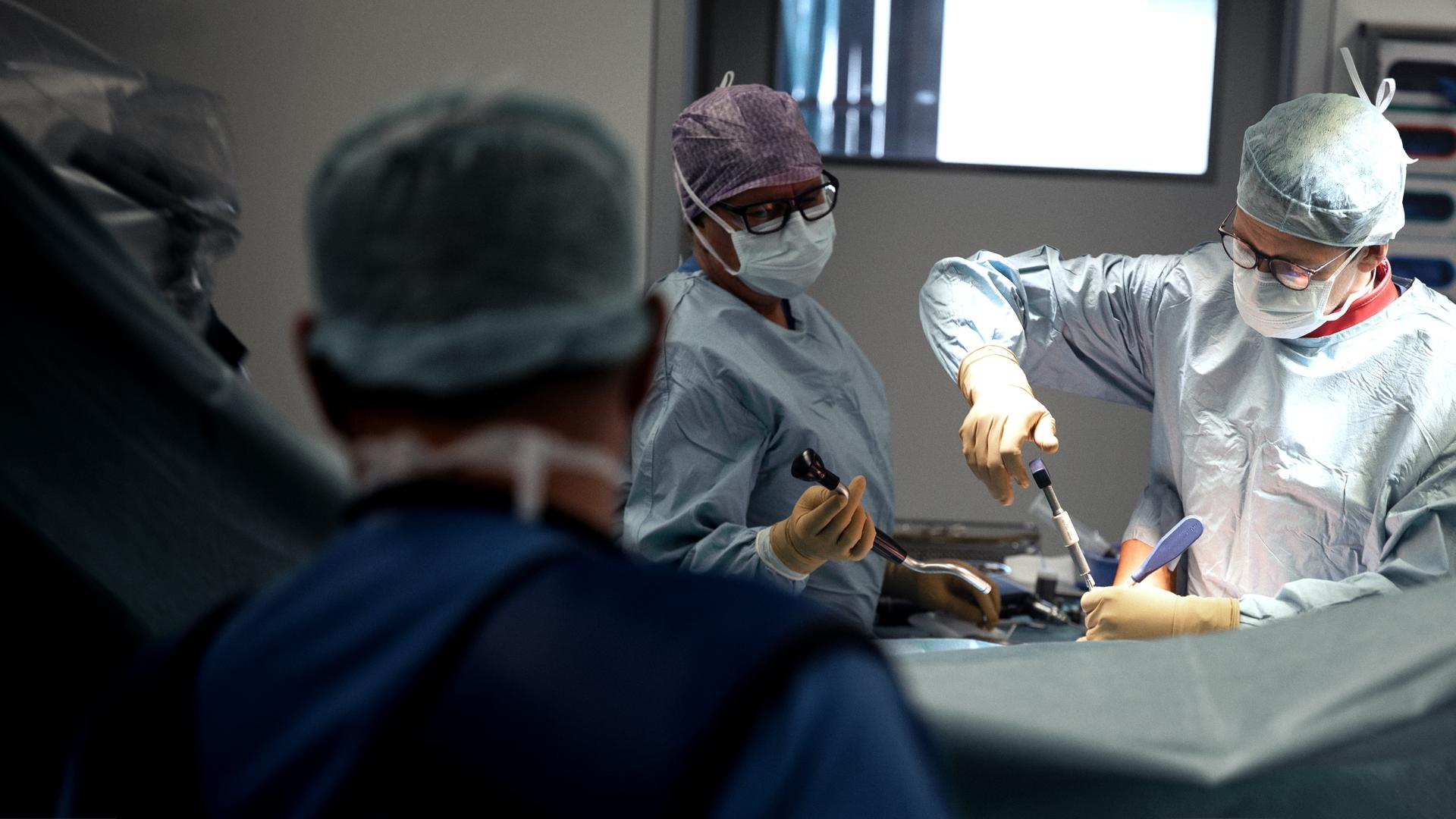 Wir sehen einen Operationssaal mit drei Chrirurgen, jeweils mit OP-Haube, Atemschutzmaske, Kittel und Handschuhe. Zwei von ihnen hantieren mit Werkzeug an einem Patienten, der im Bild nicht zu sehen ist.