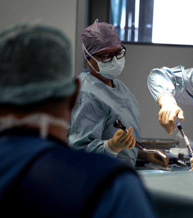 Wir sehen einen Operationssaal mit drei Chrirurgen, jeweils mit OP-Haube, Atemschutzmaske, Kittel und Handschuhe. Zwei von ihnen hantieren mit Werkzeug an einem Patienten, der im Bild nicht zu sehen ist.