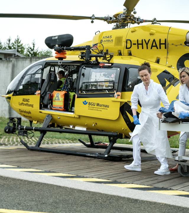 Auf dem Helikopterlandeplatz steht im Hintergrund ein Helikopter der ADAC Luftrettung/BGU Murnau. Aus dem rechten Bildrand kommen mehrere Notsanitäter mit einer Krankenliege hereingerollt.