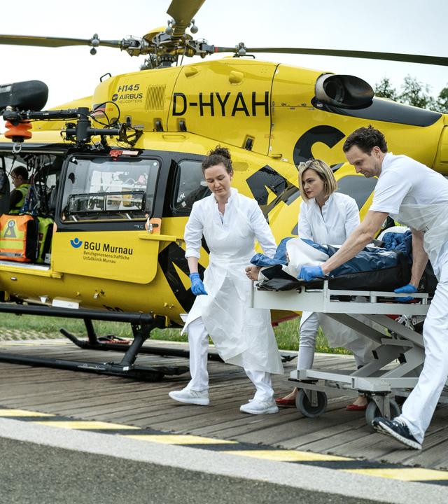Auf dem Helikopterlandeplatz steht im Hintergrund ein Helikopter der ADAC Luftrettung/BGU Murnau. Aus dem rechten Bildrand kommen mehrere Notsanitäter mit einer Krankenliege hereingerollt.