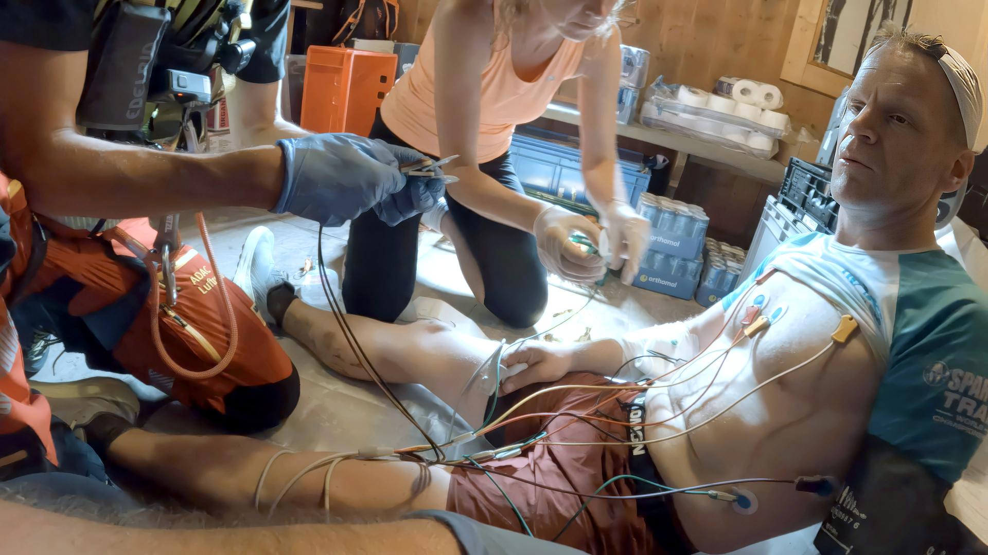 Ein Mann in Trailrunning-Outfit sitzt auf dem Boden. Sein T-Shirt ist hochgezogen, an seiner Brust hängen die Elektroden eines mobilen EKG-Geräts.