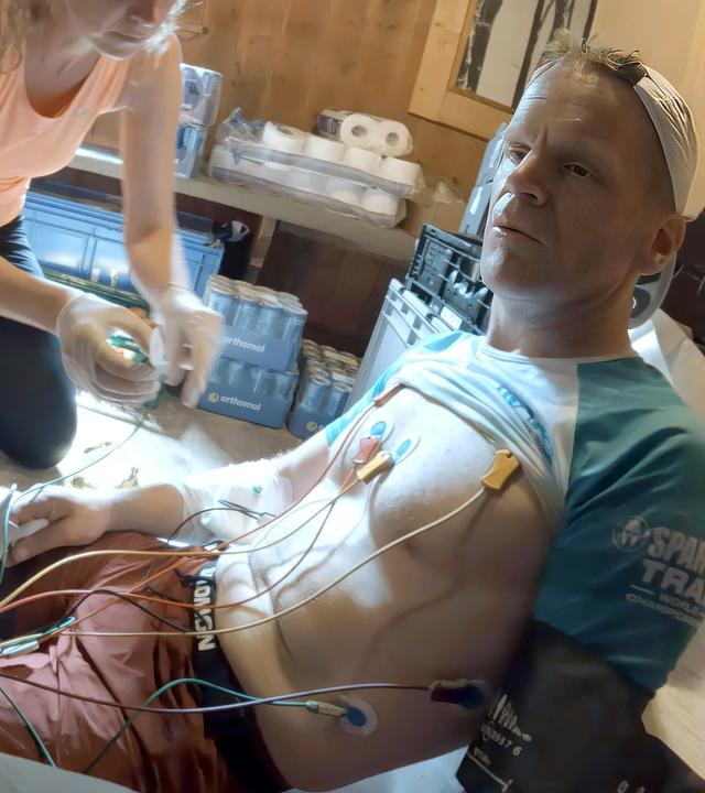 Ein Mann in Trailrunning-Outfit sitzt auf dem Boden. Sein T-Shirt ist hochgezogen, an seiner Brust hängen die Elektroden eines mobilen EKG-Geräts.