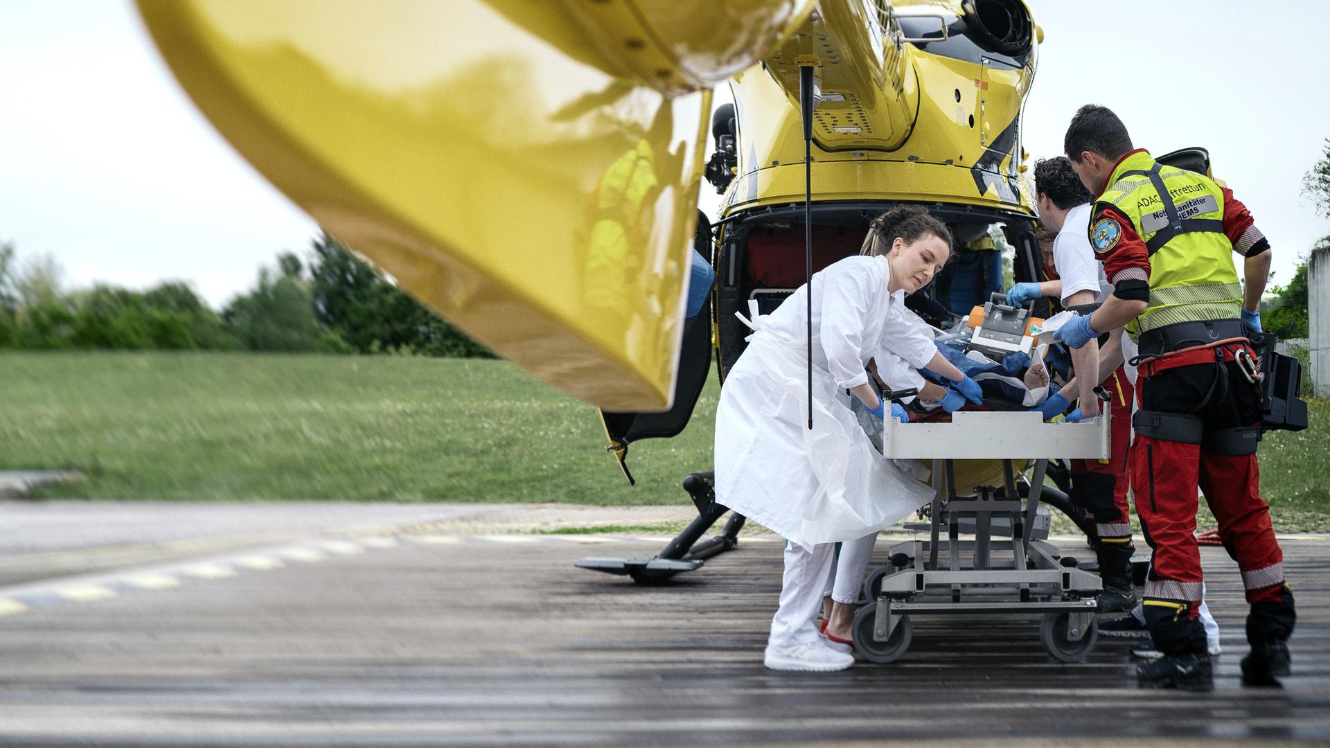Wir blicken unter dem Heck eines Nothubschraubers auf einem Landeplatz, in den ein Patient von mehreren Notfallsanitätern von einer Krankenliege geladen wird.
