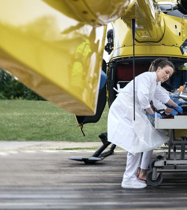 Wir blicken unter dem Heck eines Nothubschraubers auf einem Landeplatz, in den ein Patient von mehreren Notfallsanitätern von einer Krankenliege geladen wird.