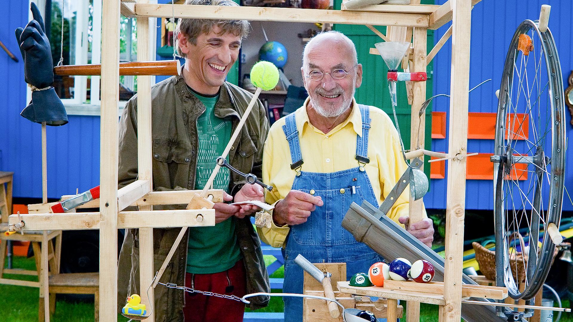Fritz Fuchs und Peter Lustig stehen an einer Erfindung und lachen.