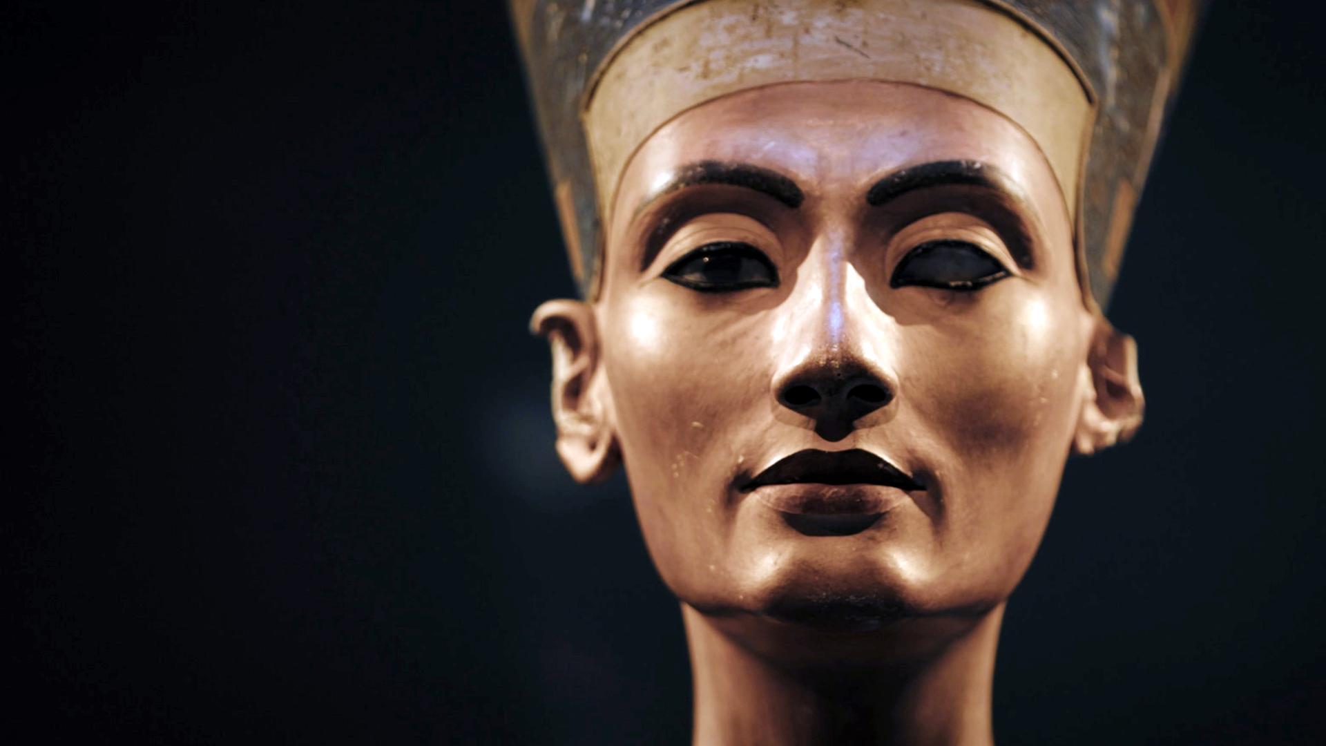 Vor schwarzem Hintergrund steht die Skulptur eines Frauenkopfes. Sie trägt eine sich nach oben erweiternde, blaue Kopfbedeckung mit einem goldenen Band über der Stirn. Auf der blauen Kopfbedeckung sind in Weiß Verzierungen zu erkennen. Das Gesicht der Frau ist ebenmäßig, ihre Augen liegen im Schatten. An einem Auge fehlt die Iris. Ein Ohr ist nicht vollständig.