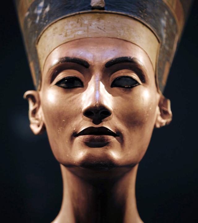 Vor schwarzem Hintergrund steht die Skulptur eines Frauenkopfes. Sie trägt eine sich nach oben erweiternde, blaue Kopfbedeckung mit einem goldenen Band über der Stirn. Auf der blauen Kopfbedeckung sind in Weiß Verzierungen zu erkennen. Das Gesicht der Frau ist ebenmäßig, ihre Augen liegen im Schatten. An einem Auge fehlt die Iris. Ein Ohr ist nicht vollständig.