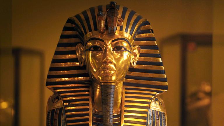 Die Totenmaske des Tutanchamun in Gold und Blau steht vor einem ockerfarbenen Hintergrund. Der Kopf wird von einem Nemes-Kopftuch bedeckt. In Stirnhöhe sitzen ein Geierkopf und der Oberkörper eine Kobra. Die Ohrläppchen sind durchstochen. Die hellen Augen sind blau umrandet, die Augenbrauen sind in blau ausgearbeitet. Am Kinn sitzt ein langer, geflochtener Zeremonialbart. An den beiden Schultern sind die Ränder eines breiten Halskragens zu sehen.