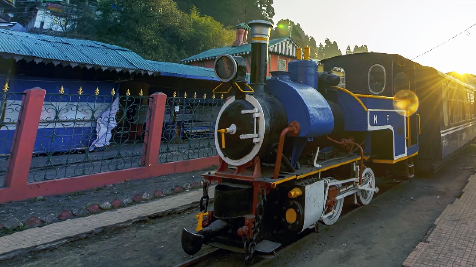 Eine kleine Dampflock mit einem blau angemalten Kessel steht auf einem gerade verlaufenden Gleis bei Sonnenuntergang. Links vom Zug steht ein Zaun, dahinter einige flache Gebäude. Rechts stehen einige Bänke und Bäume.