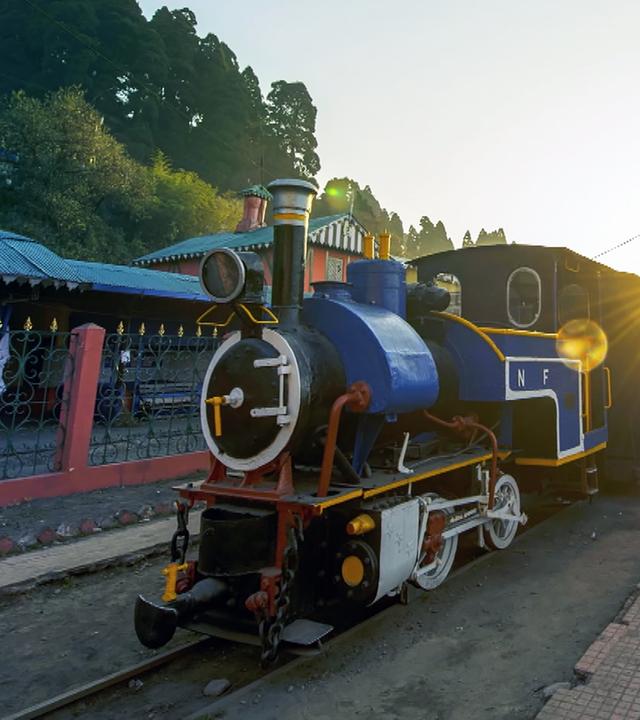 Eine kleine Dampflock mit einem blau angemalten Kessel steht auf einem gerade verlaufenden Gleis bei Sonnenuntergang. Links vom Zug steht ein Zaun, dahinter einige flache Gebäude. Rechts stehen einige Bänke und Bäume.