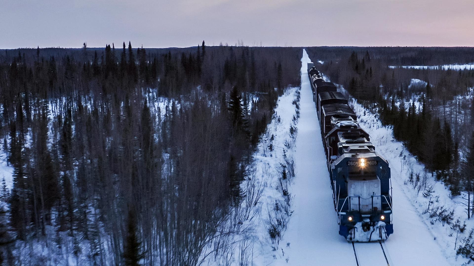 Panoramaaufnahme: Ein Güterzug durchquert eine Schneelandschaft mit kargen Bäumen bei Sonnenuntergang.