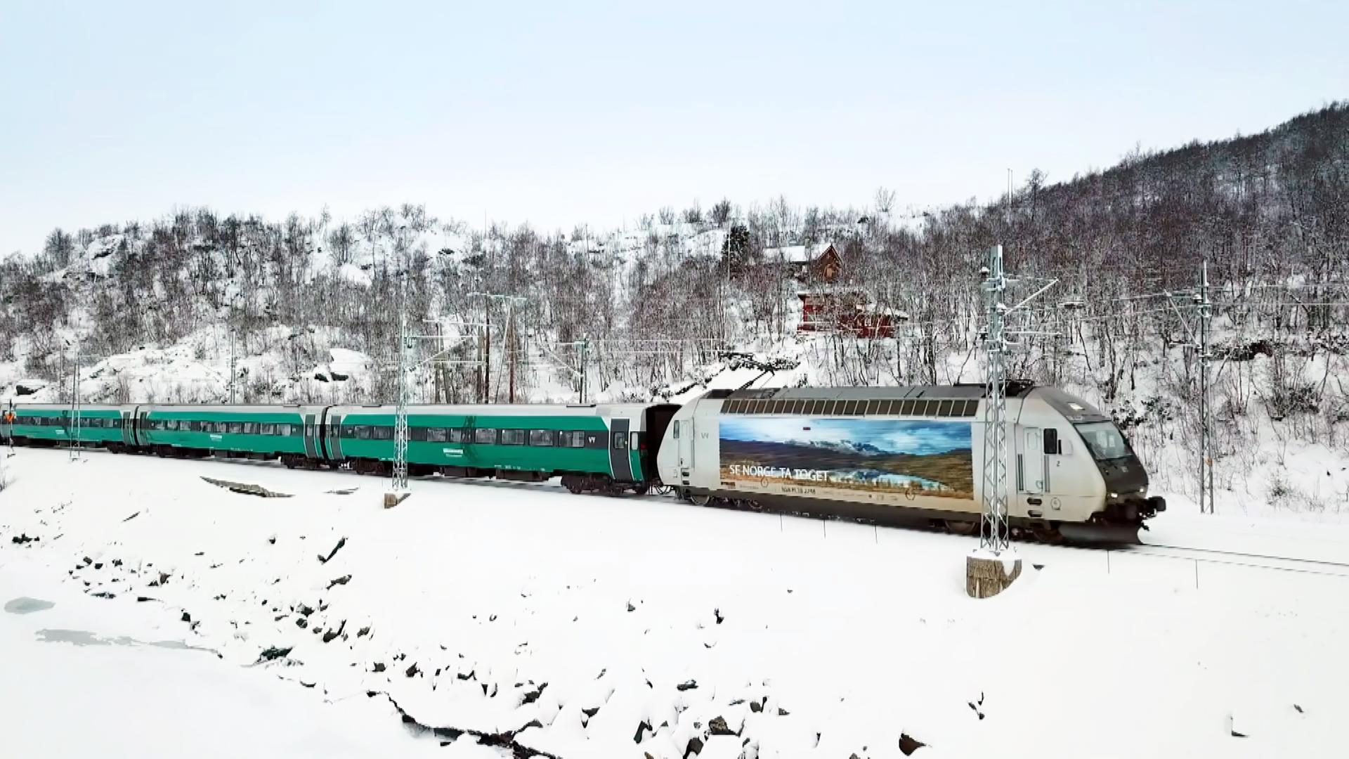 Ein Zug mit grünen Waggons und einem weißen Triebwagen, auf dem ein Bild von einer norwegischen Berglandschaft abgebildet ist, fährt durch eine schneebedeckte Landschaft.
