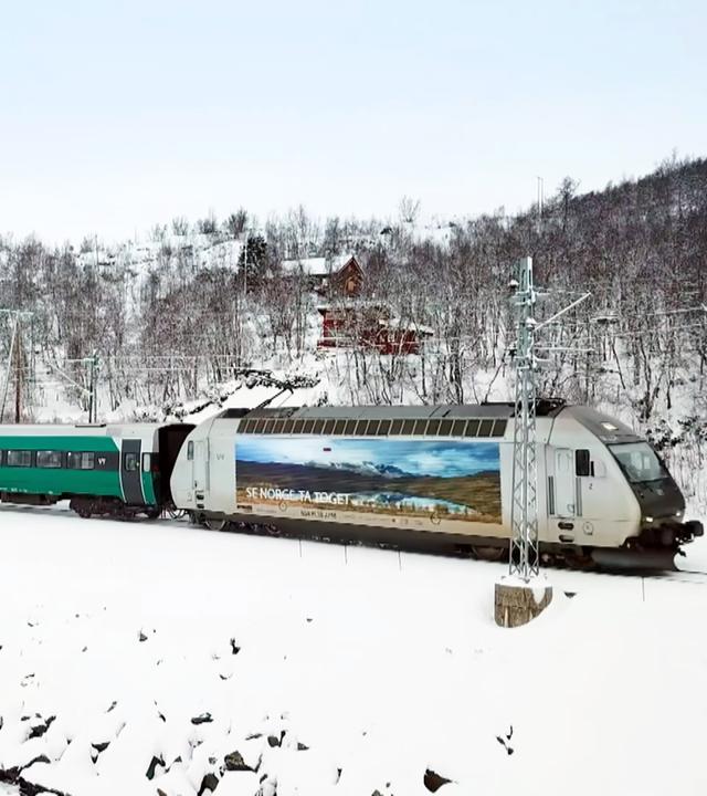 Ein Zug mit grünen Waggons und einem weißen Triebwagen, auf dem ein Bild von einer norwegischen Berglandschaft abgebildet ist, fährt durch eine schneebedeckte Landschaft.
