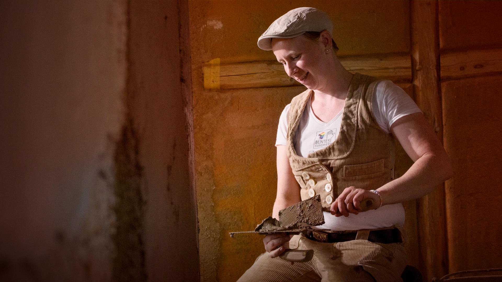 Eine Frau mit brauner Kordweste und Kappe kniet auf dem Boden vor unverputzten Wänden. Sie hat zwei Spachtel in den Händen, von links fällt Licht in die Szene.
