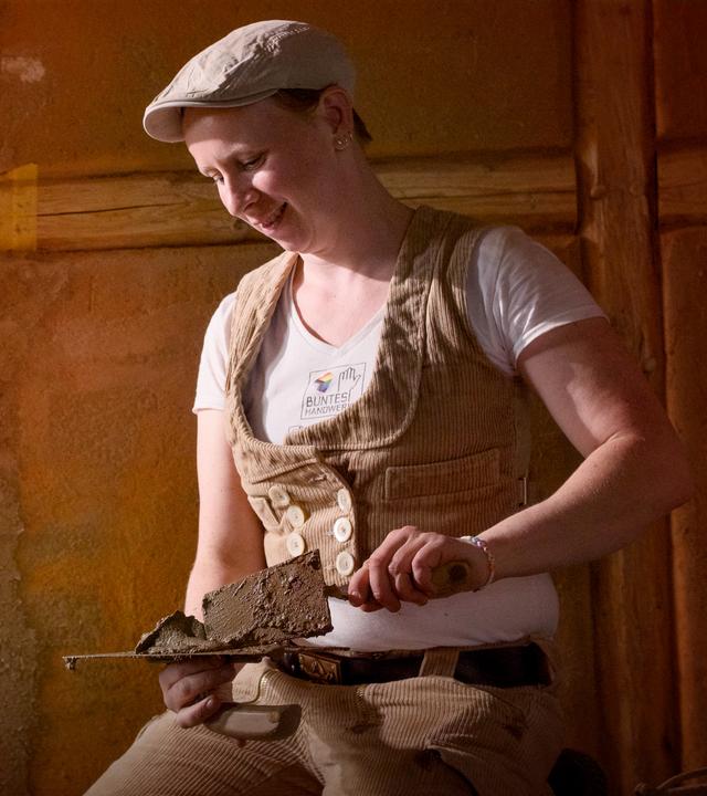 Eine Frau mit brauner Kordweste und Kappe kniet auf dem Boden vor unverputzten Wänden. Sie hat zwei Spachtel in den Händen, von links fällt Licht in die Szene.