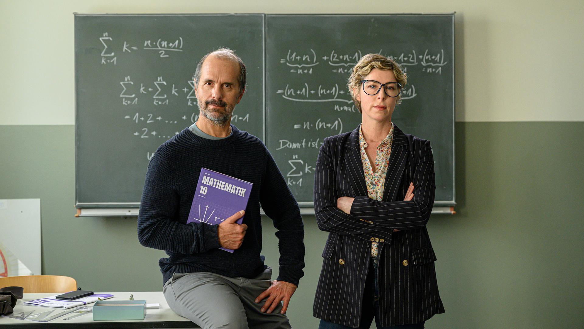 "Lehrer kann jeder!": Richard Glossat (Christoph Maria Herbst) sitzt in einem Klassenzimmer auf dem Lehrerpult und hält ein Mathematik-Buch in der Hand. Neben ihm steht Anke Glossat (Brigitte Zeh) mit verschränkten Armen und genervtem Blick. Im Hintergrund eine Schultafel, an die mit Kreide Mathematik-Formeln geschrieben stehen.