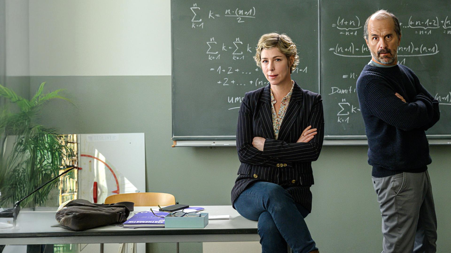 "Lehrer kann jeder!": Richard Glossat (Christoph Maria Herbst) sitzt in einem Klassenzimmer auf dem Lehrerpult und hält ein Mathematik-Buch in der Hand. Neben ihm steht Anke Glossat (Brigitte Zeh) mit verschränkten Armen und genervtem Blick. Im Hintergrund eine Schultafel, an die mit Kreide Mathematik-Formeln geschrieben stehen.