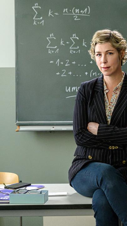 "Lehrer kann jeder!": Richard Glossat (Christoph Maria Herbst) sitzt in einem Klassenzimmer auf dem Lehrerpult und hält ein Mathematik-Buch in der Hand. Neben ihm steht Anke Glossat (Brigitte Zeh) mit verschränkten Armen und genervtem Blick. Im Hintergrund eine Schultafel, an die mit Kreide Mathematik-Formeln geschrieben stehen.