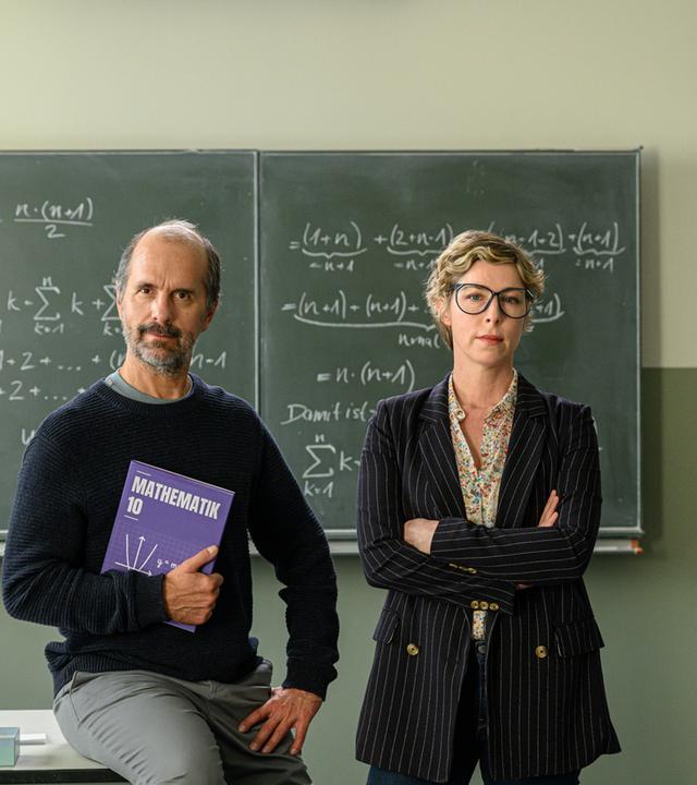 "Lehrer kann jeder!": Richard Glossat (Christoph Maria Herbst) sitzt in einem Klassenzimmer auf dem Lehrerpult und hält ein Mathematik-Buch in der Hand. Neben ihm steht Anke Glossat (Brigitte Zeh) mit verschränkten Armen und genervtem Blick. Im Hintergrund eine Schultafel, an die mit Kreide Mathematik-Formeln geschrieben stehen.