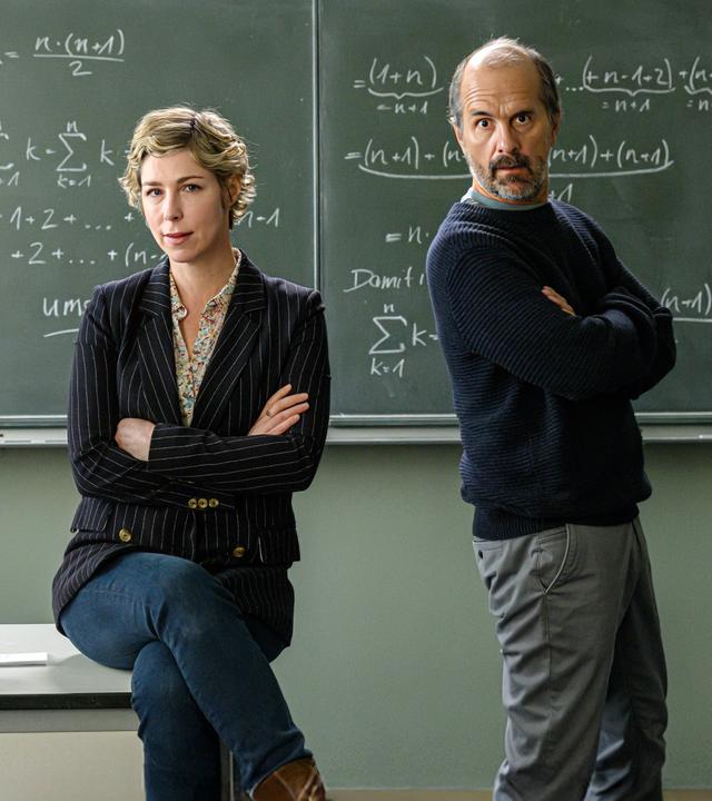 "Lehrer kann jeder!": Richard Glossat (Christoph Maria Herbst) sitzt in einem Klassenzimmer auf dem Lehrerpult und hält ein Mathematik-Buch in der Hand. Neben ihm steht Anke Glossat (Brigitte Zeh) mit verschränkten Armen und genervtem Blick. Im Hintergrund eine Schultafel, an die mit Kreide Mathematik-Formeln geschrieben stehen.