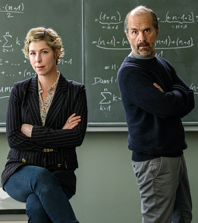 "Lehrer kann jeder!": Richard Glossat (Christoph Maria Herbst) sitzt in einem Klassenzimmer auf dem Lehrerpult und hält ein Mathematik-Buch in der Hand. Neben ihm steht Anke Glossat (Brigitte Zeh) mit verschränkten Armen und genervtem Blick. Im Hintergrund eine Schultafel, an die mit Kreide Mathematik-Formeln geschrieben stehen.