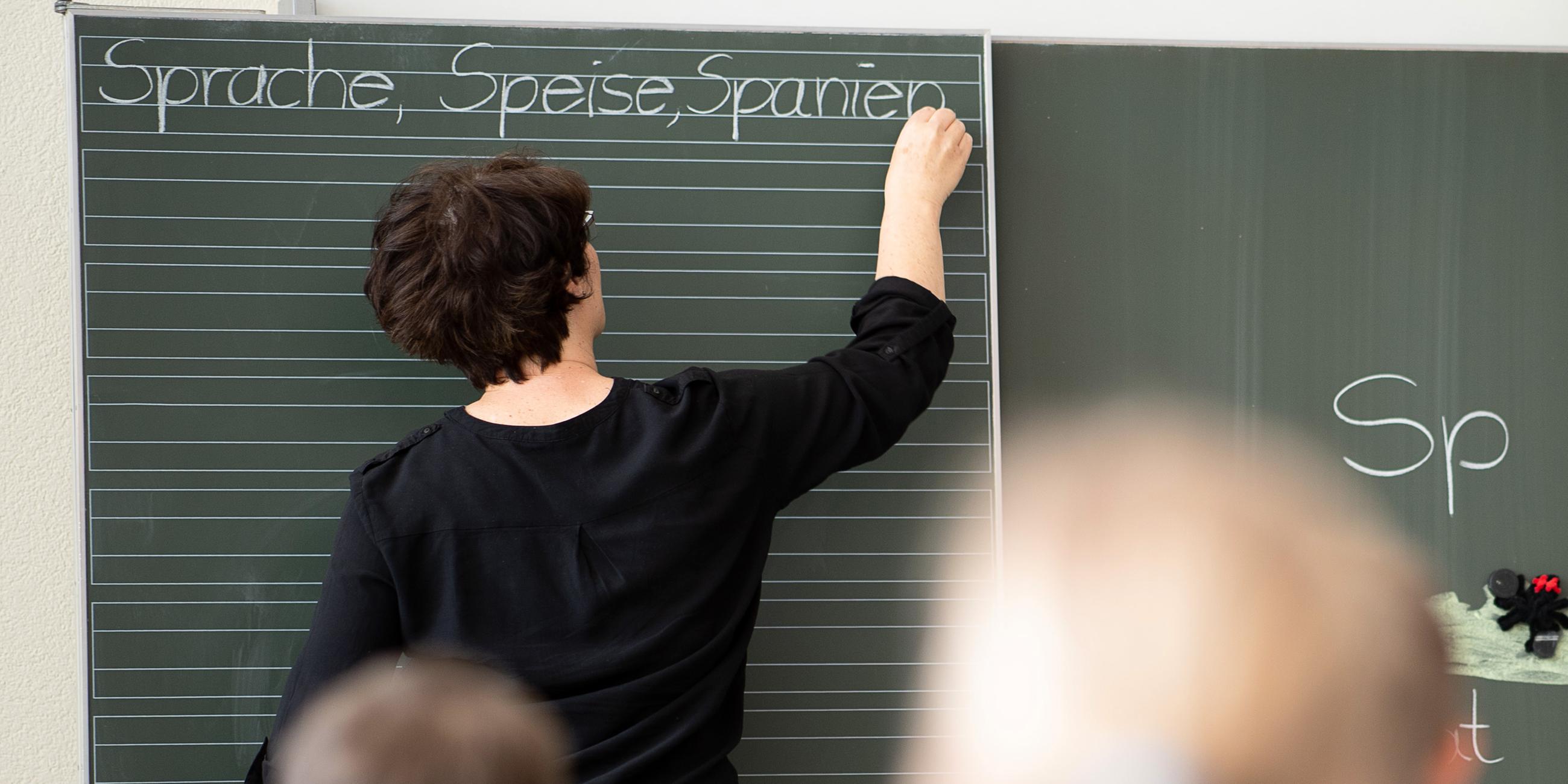Eine Lehrerin steht in einem vollen Klassenzimmer und schreibt etwas an die Tafel.