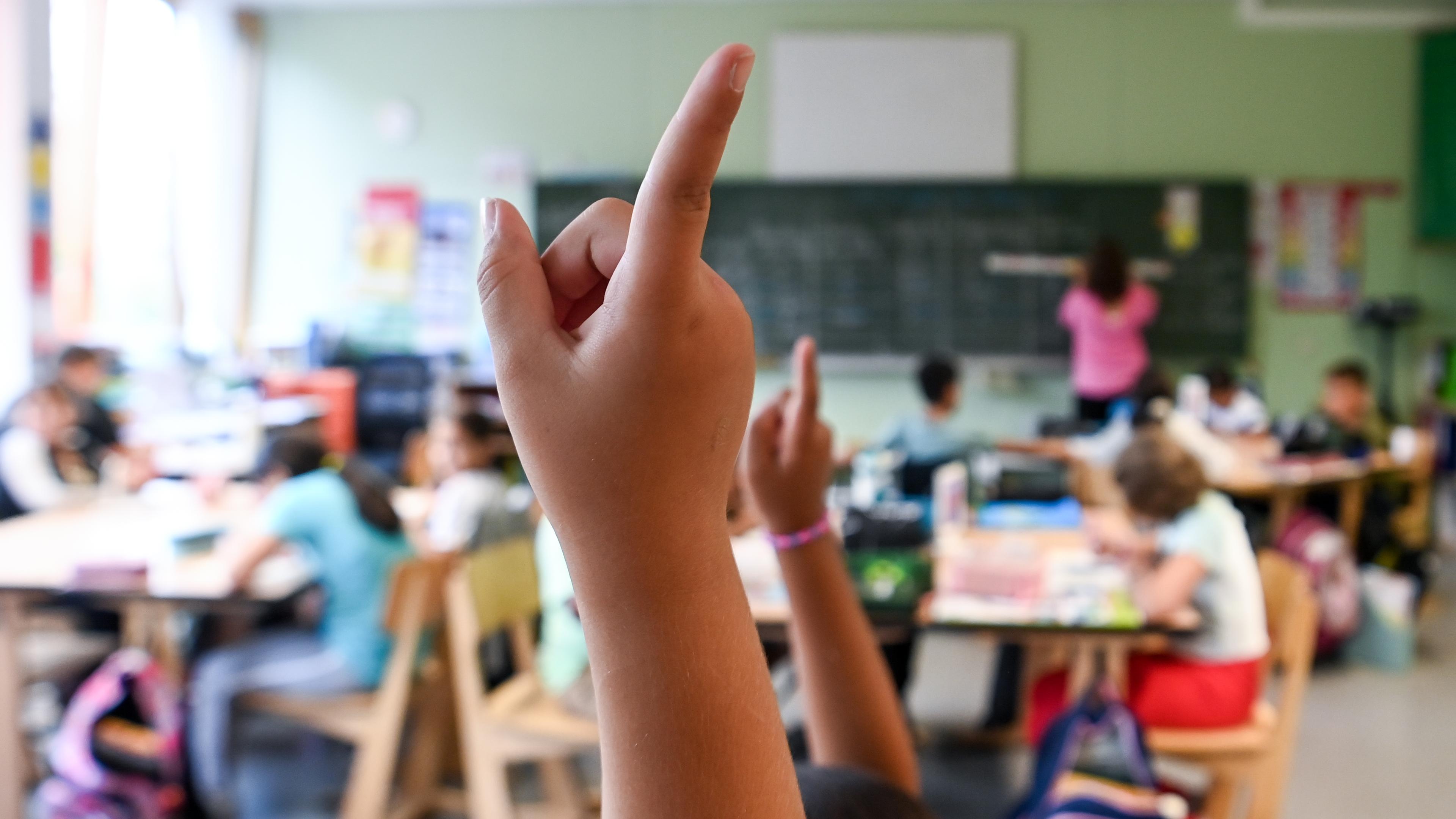 Ein volles Klassenzimmer von hinten, ein Kind hebt die Hand.