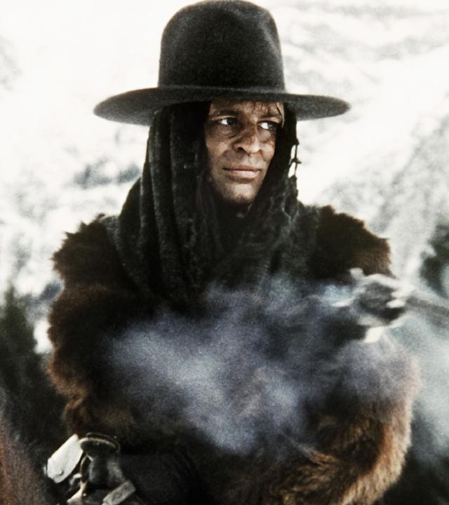 Loco (Klaus Kinski) sitzt inmitten einer verschneiten Berglandschaft auf einem Pferd und trägt einen dunklen Hut sowie einen dicken Pelzmantel. Dabei feuert er mit seinem Gewehr, aus dessen Mündung Rauch aufsteigt.