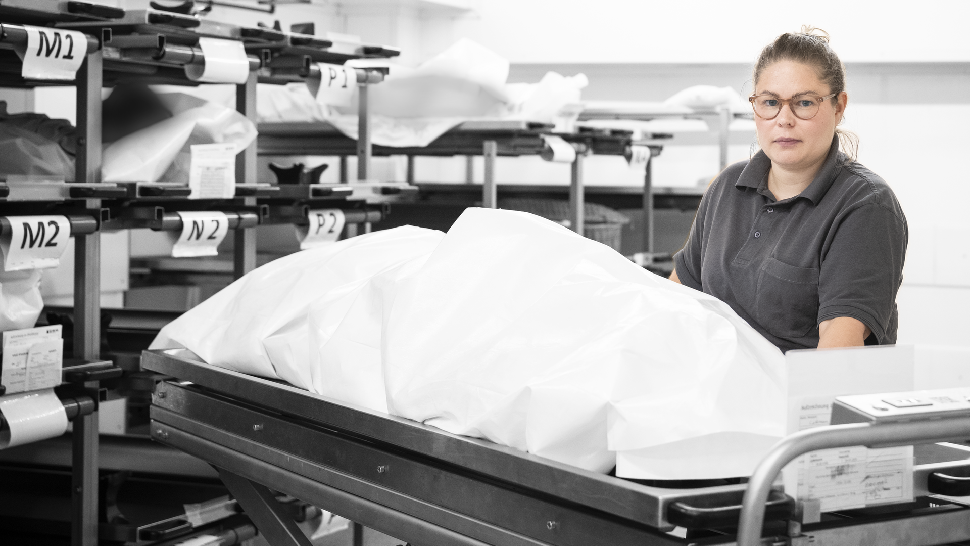 Marleen steht vor einer Pritsche, auf der ein Tuch über einer Leiche liegt.