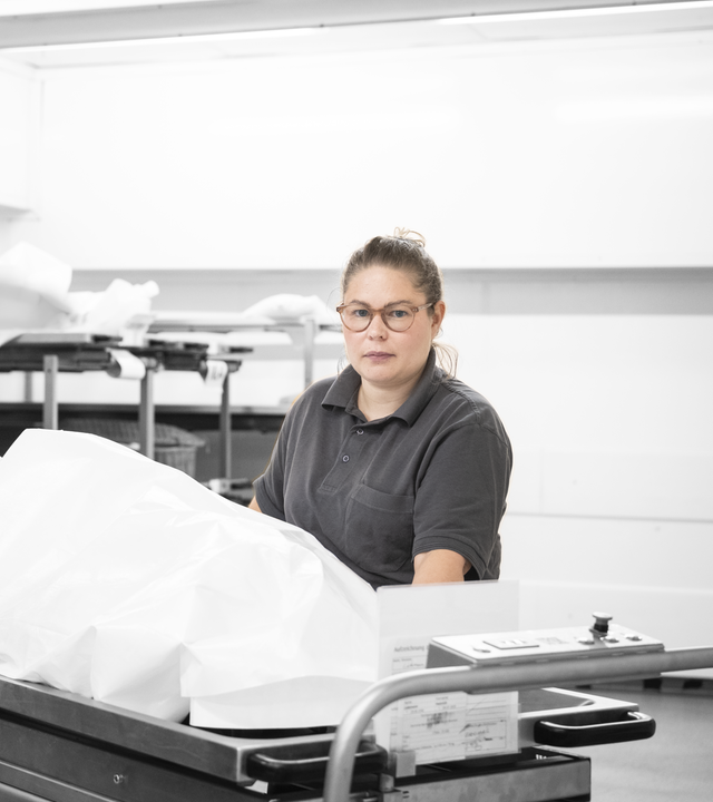 Marleen steht vor einer Pritsche, auf der ein Tuch über einer Leiche liegt.