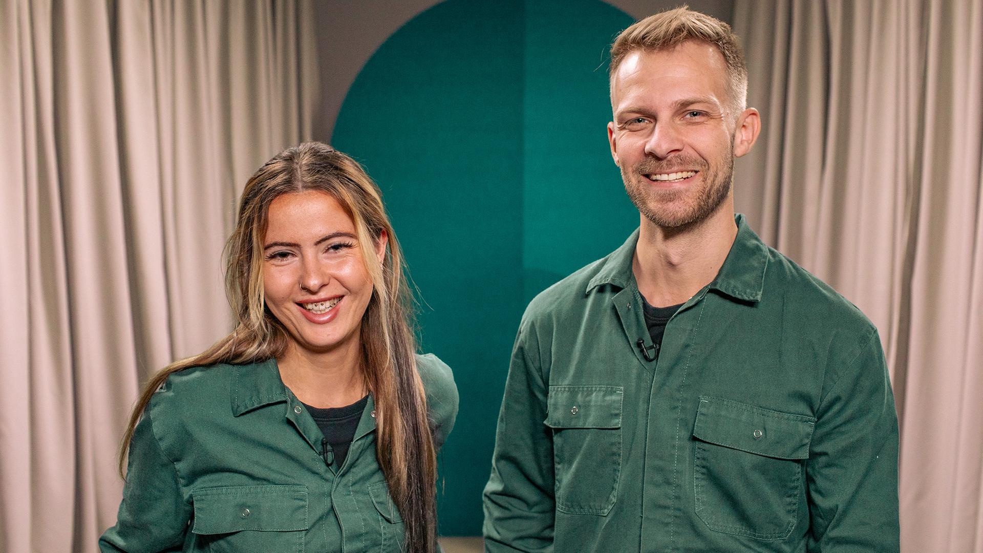Die beiden Gäste der Folge, eine Frau und ein Mann, tragen den gleichen grünen Overall und blicken in die Kamera.