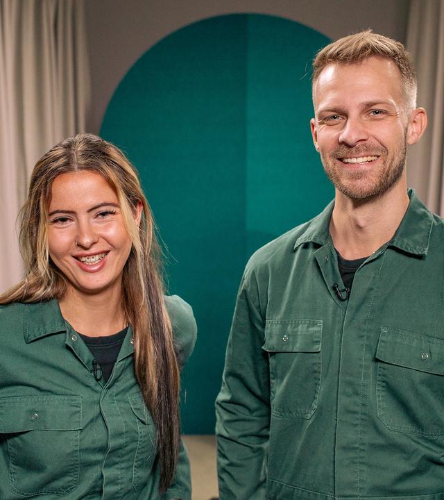 Die beiden Gäste der Folge, eine Frau und ein Mann, tragen den gleichen grünen Overall und blicken in die Kamera.