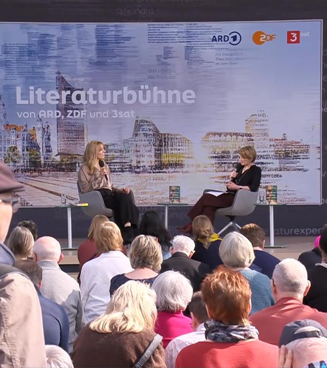 Zwei Frauen sitzen auf einer Bühne vor Publikum und unterhalten sich. Im Hintergrund ist ein Bild eingeblendet, auf dem Lesebühne steht.