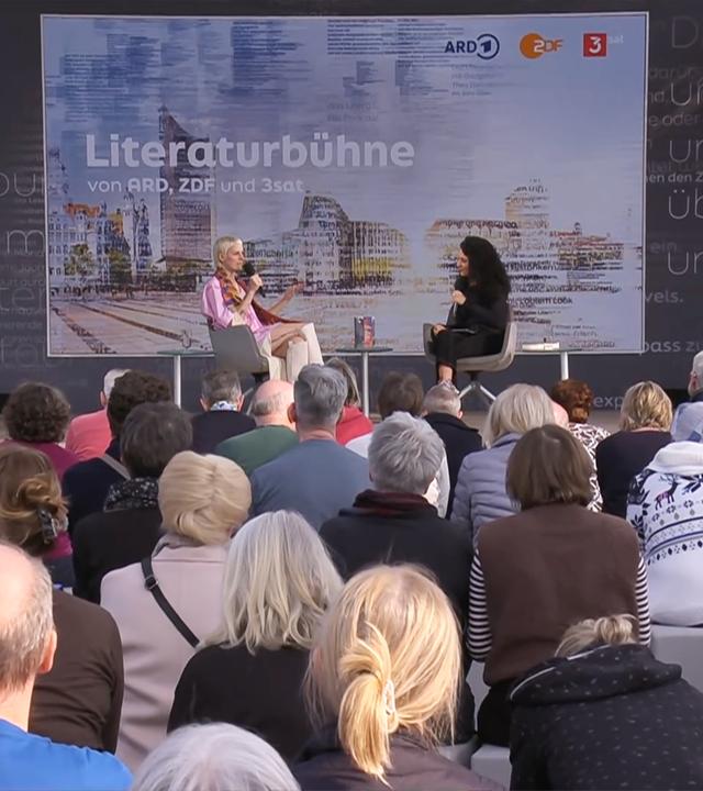 Zwei Frauen sitzen auf einer Bühne vor Publikum und unterhalten sich. Im Hintergrund ist ein Bild eingeblendet, auf dem Lesebühne steht.