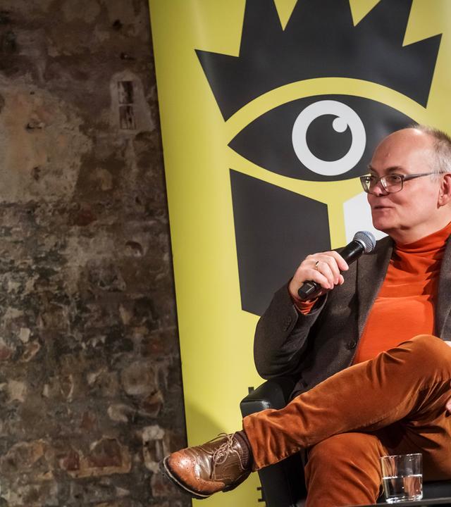 Ein Mann mittleren Alters sitzt auf einem Podium mit dem Logo der Leipziger Buchmesse im Hitergrund.