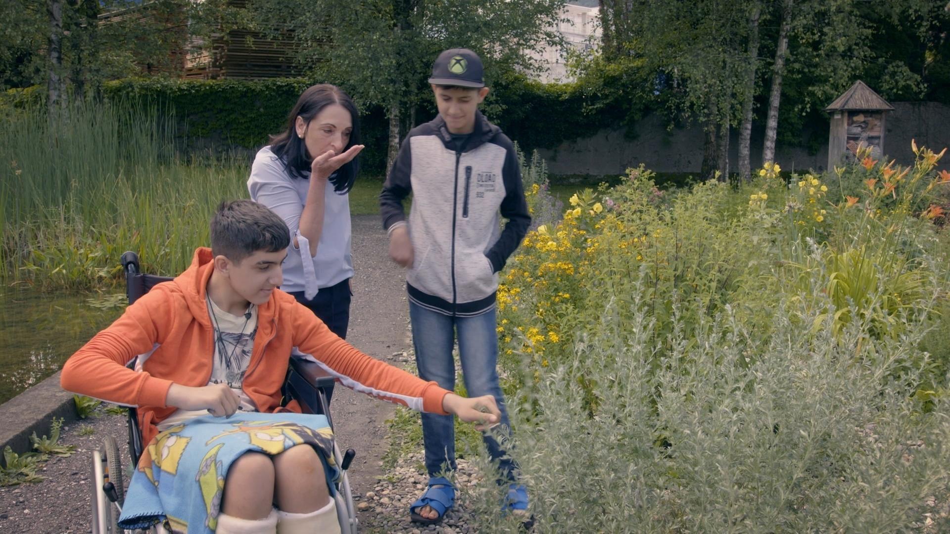 Heidi Ludescher mit zwei Jungs in einem Park mit Wildblumen. Ein Junge mit rosa Jacke sitzt im Rollstuhl.