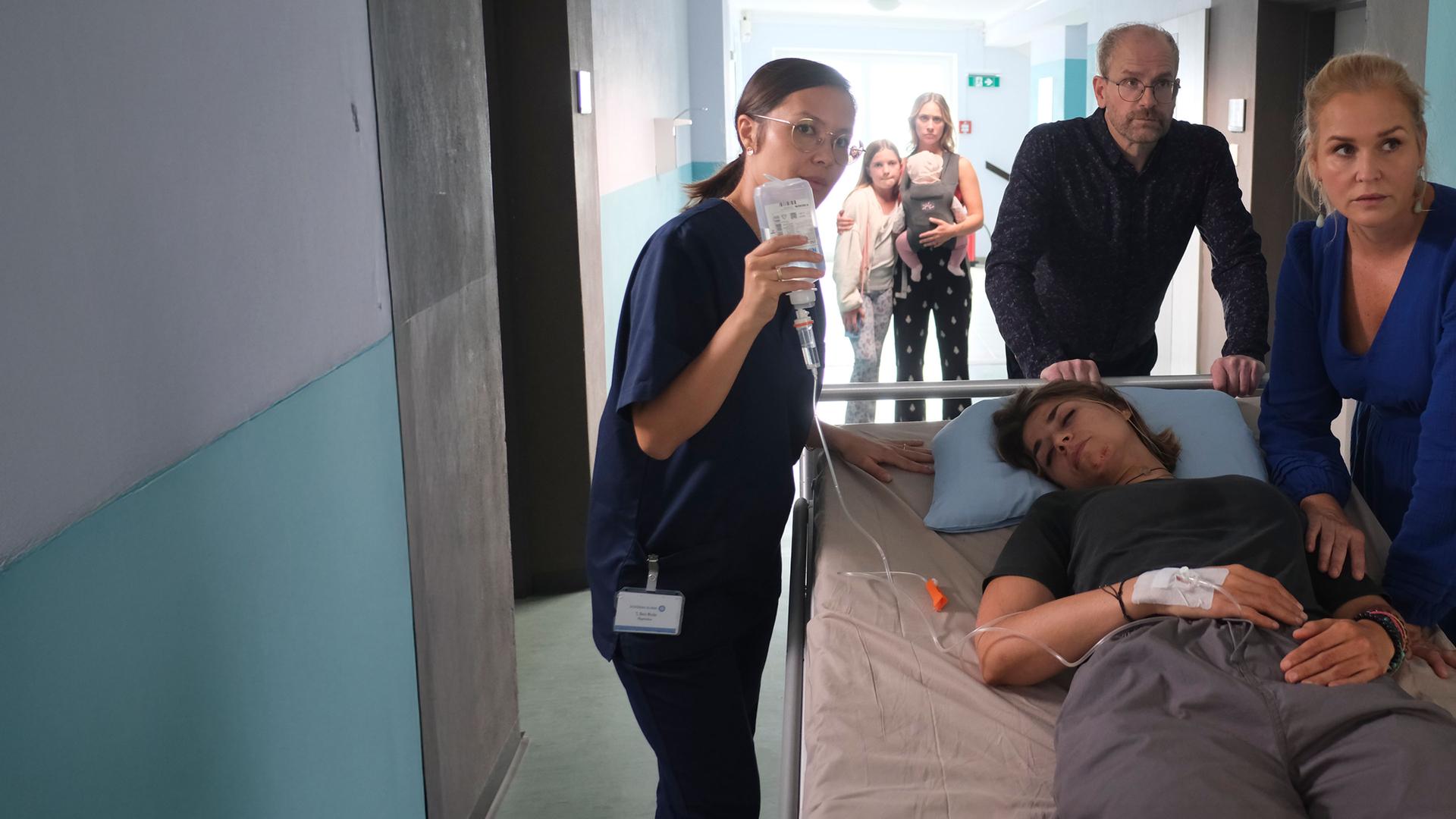 "Lena Lorenz - Blinder Passagier": Dr. Stefan Keller (Thomas Limpinsel) und Lena Lorenz (Judith Hoersch) schieben ein Krankenbett, auf dem Enissa Heydari (Jaëla Probst) liegt, durch einen Krankenhausflur zur Intensivstation. Eine Pflegerin (Komparsin) geht nebenher und hält eine Infusionsflasche, deren Nadel an Enissas Handrücken angebracht ist, in die Höhe. Im Hintergrund steht Mina Heydari (Sina Tkotsch) mit Baby Ella und Tochter Lara (Martha Felipa Abel).
