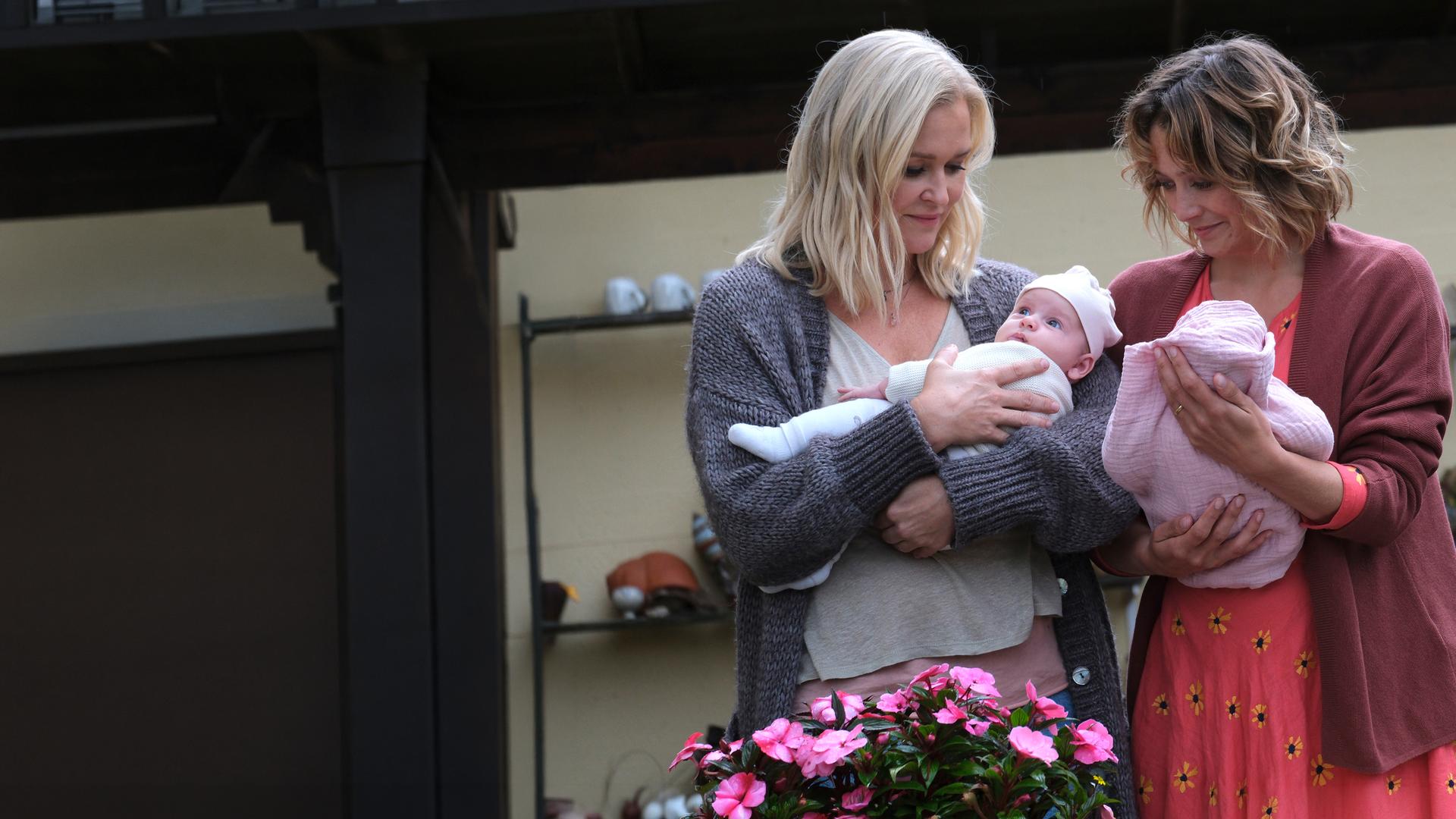"Lena Lorenz - Das halbe Lottchen": Auf einer Terrasse stehen Lena Lorenz (Judith Hoersch) und Magali Bernbauer (Hanna Plaß) nebeneinander. Jede hält eines der beiden Zwillingsmädchen im Arm.