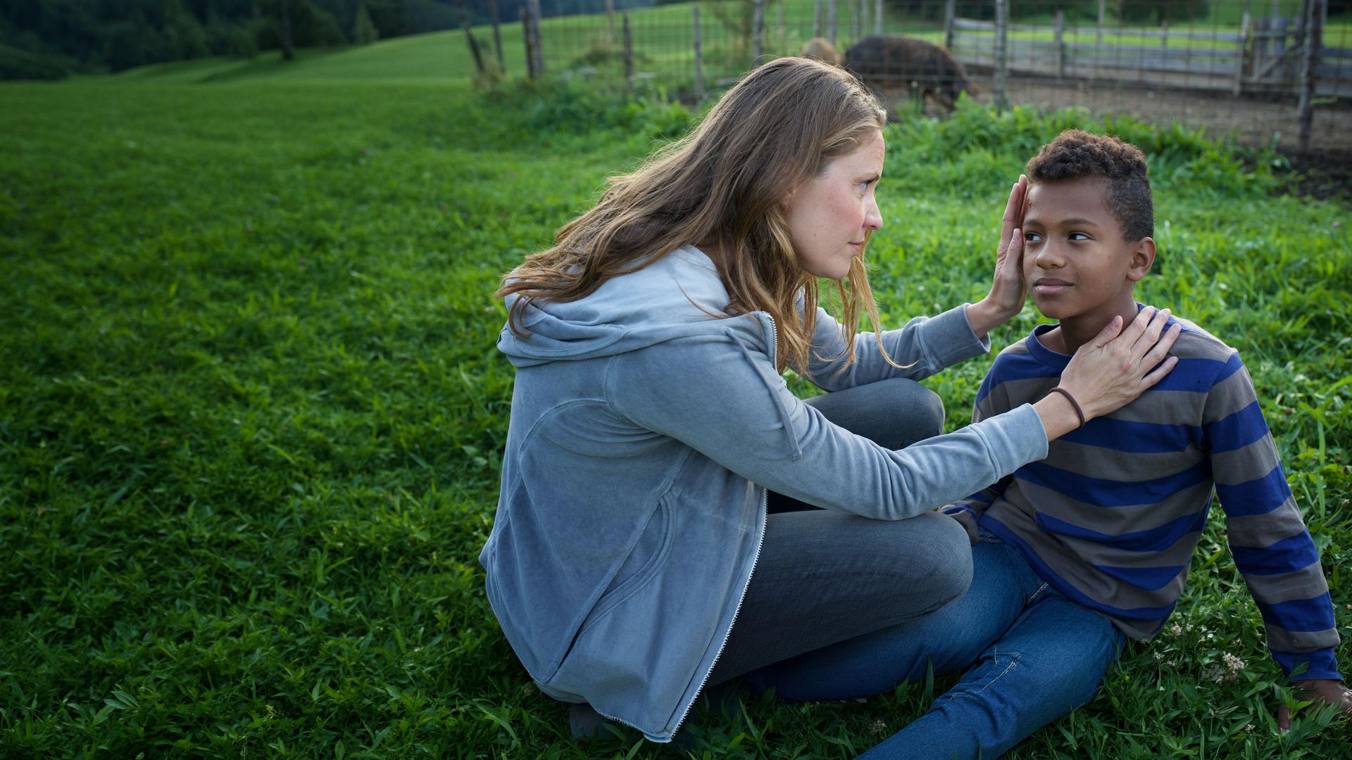 "Lena Lorenz - Von weit her": Lena Lorenz (Patricia Aulitzky) sitzt im Gras in der Hocke neben Anbessa (Ruben Felipe) und schaut sich dessen Schürfwunde an der Stirn an.
