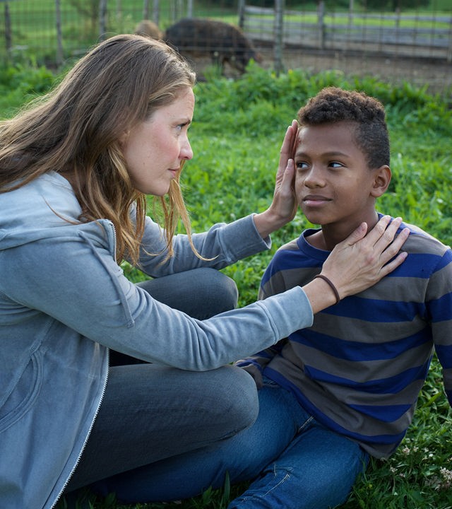"Lena Lorenz - Von weit her": Lena Lorenz (Patricia Aulitzky) sitzt im Gras in der Hocke neben Anbessa (Ruben Felipe) und schaut sich dessen Schürfwunde an der Stirn an.