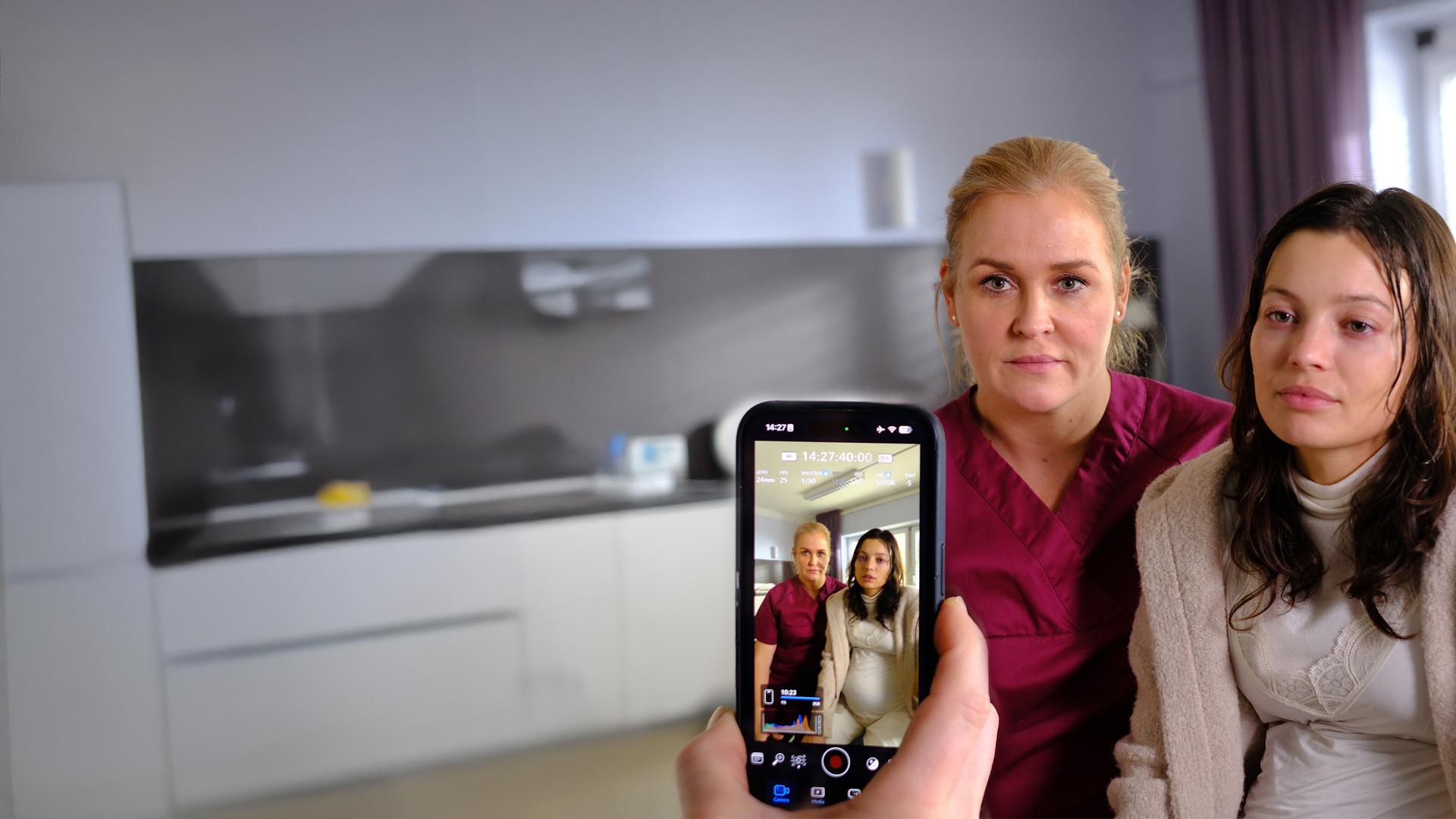 "Lena Lorenz - Vor aller Augen": In einem Krankenzimmer sitzen Lena Lorenz (Judith Hoersch) und Nadja Brettschneider (Vivien König) nebeneinander auf einem Bett. Beide schauen in eine Handykamera, die auf sie gerichtet wird.