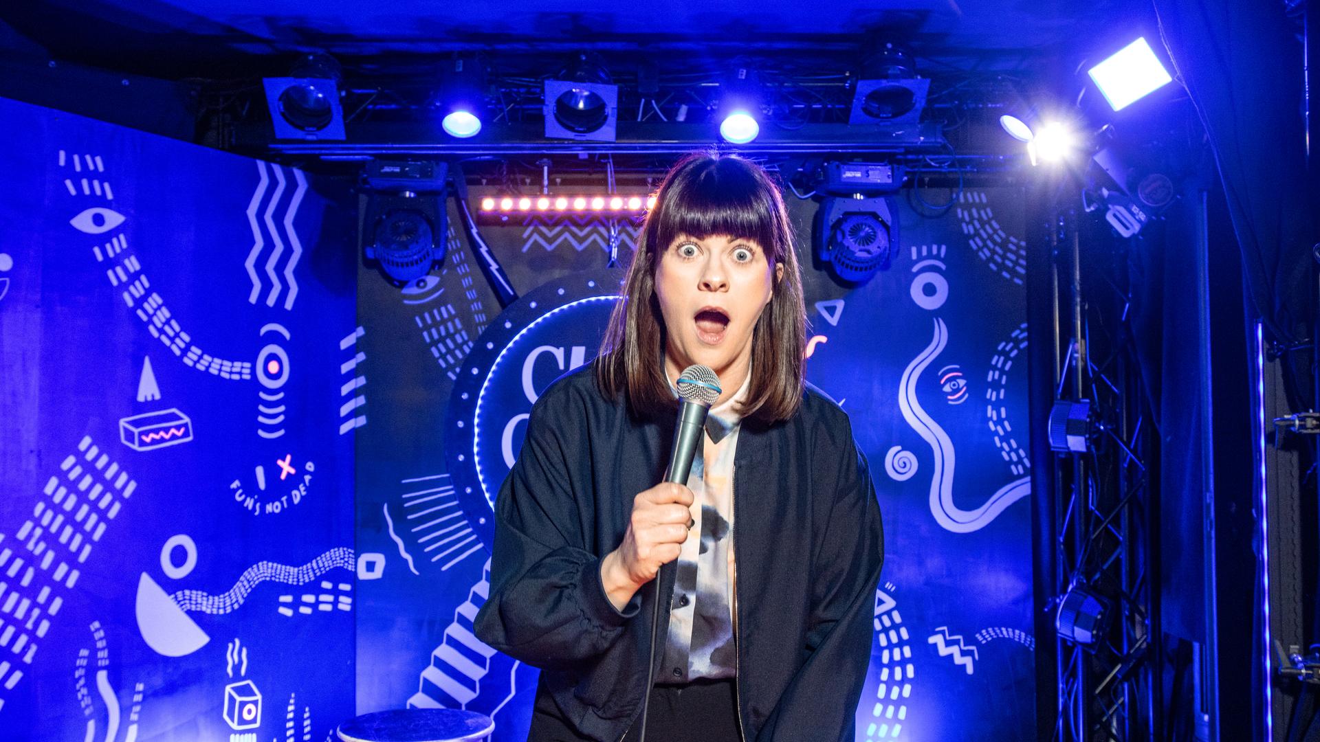 Lena Loster steht auf der Bühne und hält ein Mikrofon in ihrer rechten Hand. Der Hintergrund ist blau beleuchtet. 