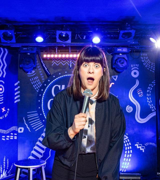 Lena Loster steht auf der Bühne und hält ein Mikrofon in ihrer rechten Hand. Der Hintergrund ist blau beleuchtet. 