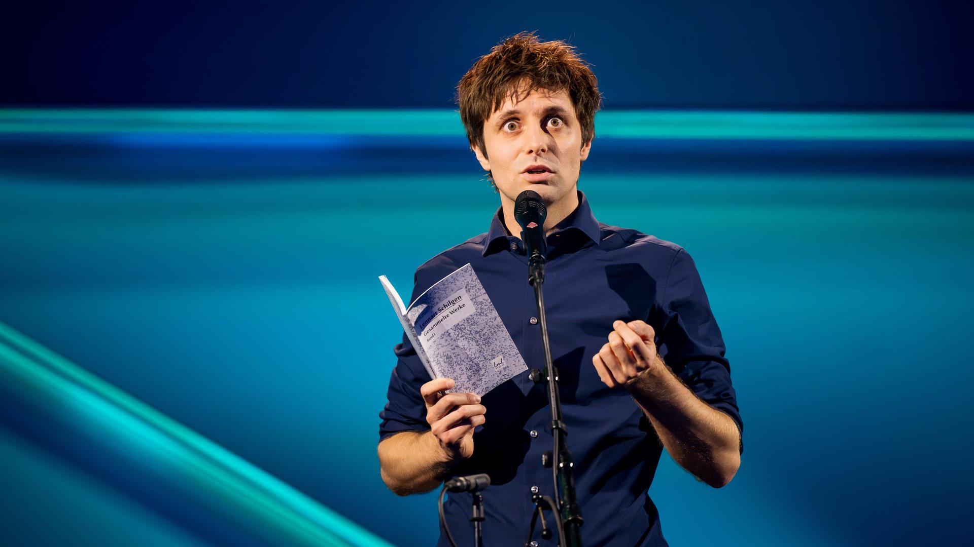 Lennart Schilgen steht in der Mitte des Bildes. In seiner rechten Hand hält er ein Buch. Vor ihm steht ein Mikrofon. Der Hintergrund ist blau beleuchtet.