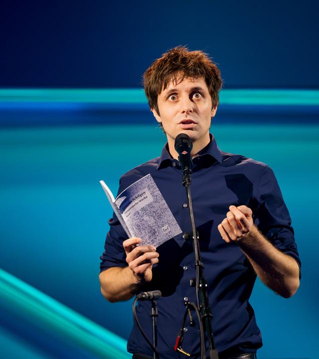 Lennart Schilgen steht in der Mitte des Bildes. In seiner rechten Hand hält er ein Buch. Vor ihm steht ein Mikrofon. Der Hintergrund ist blau beleuchtet.