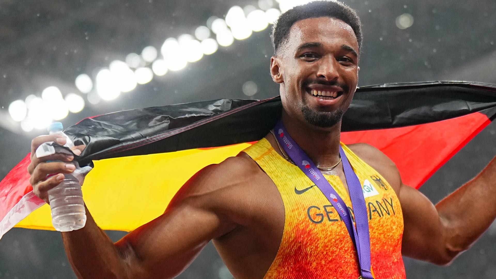 Leo Neugebauer ist Zehnkampf-Weltmeister. Er posiert mit einer Deutschlandflagge und schaut lächelnd in die Kamera.