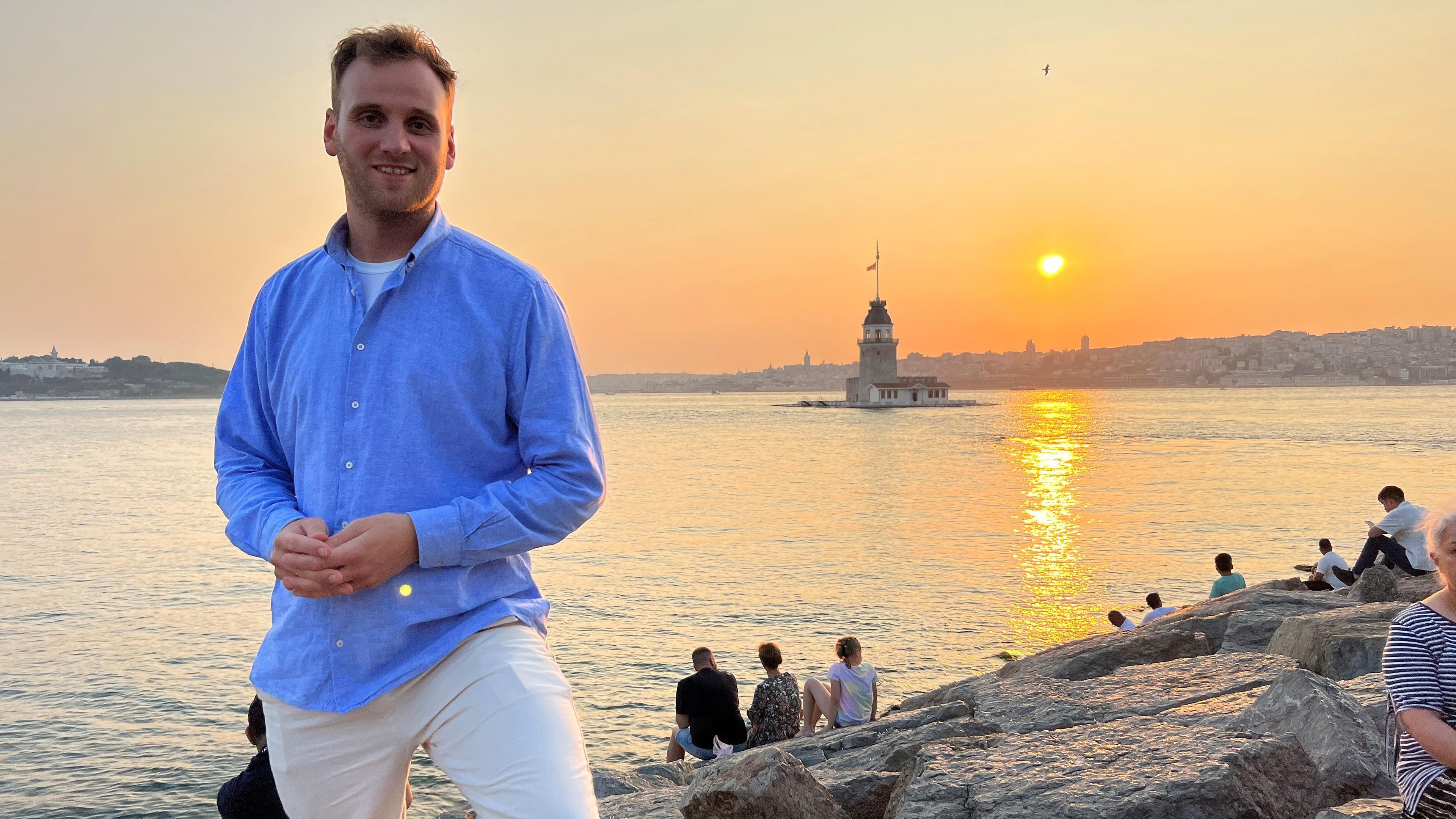 Leon Windscheid steht bei Sonnenuntergang am Bosporus und schaut in die Kamera. Hinter ihm sitzen mehrere Personen auf Felsen und schauen auf das Wasser.