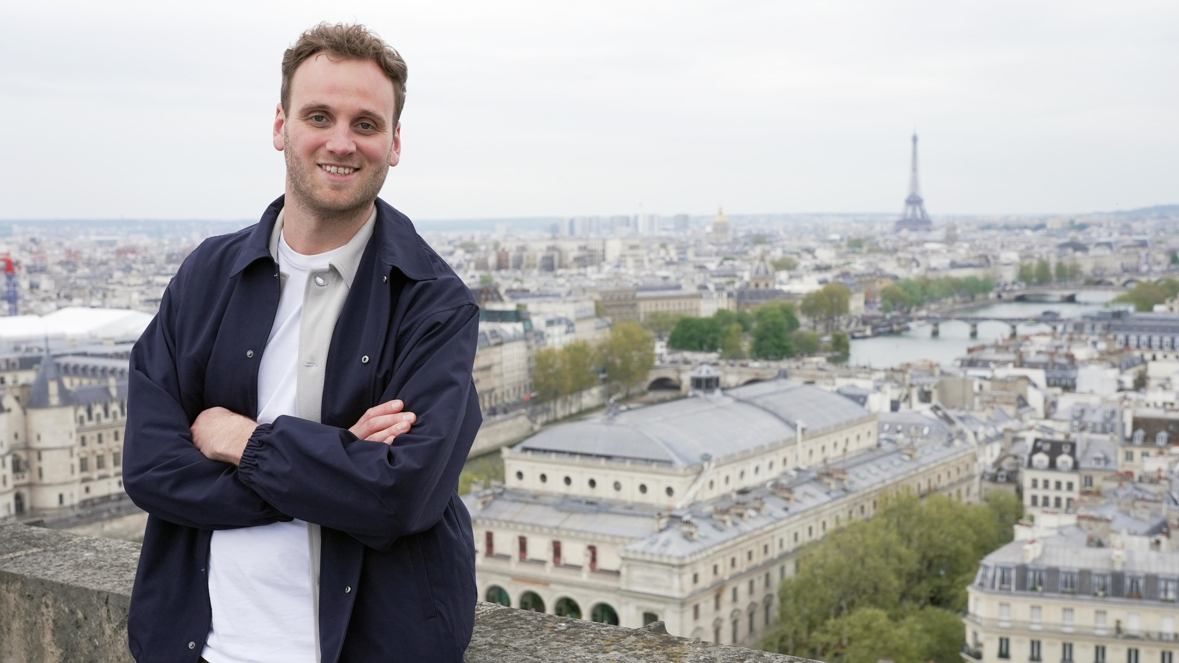 Moderator Leon Windscheid steht auf einem Balkon in Paris, hinter ihm liegt die Stadt mit dem Eiffelturm. Er hat die Arme verschränkt und schaut lächelnd in die Kamera.