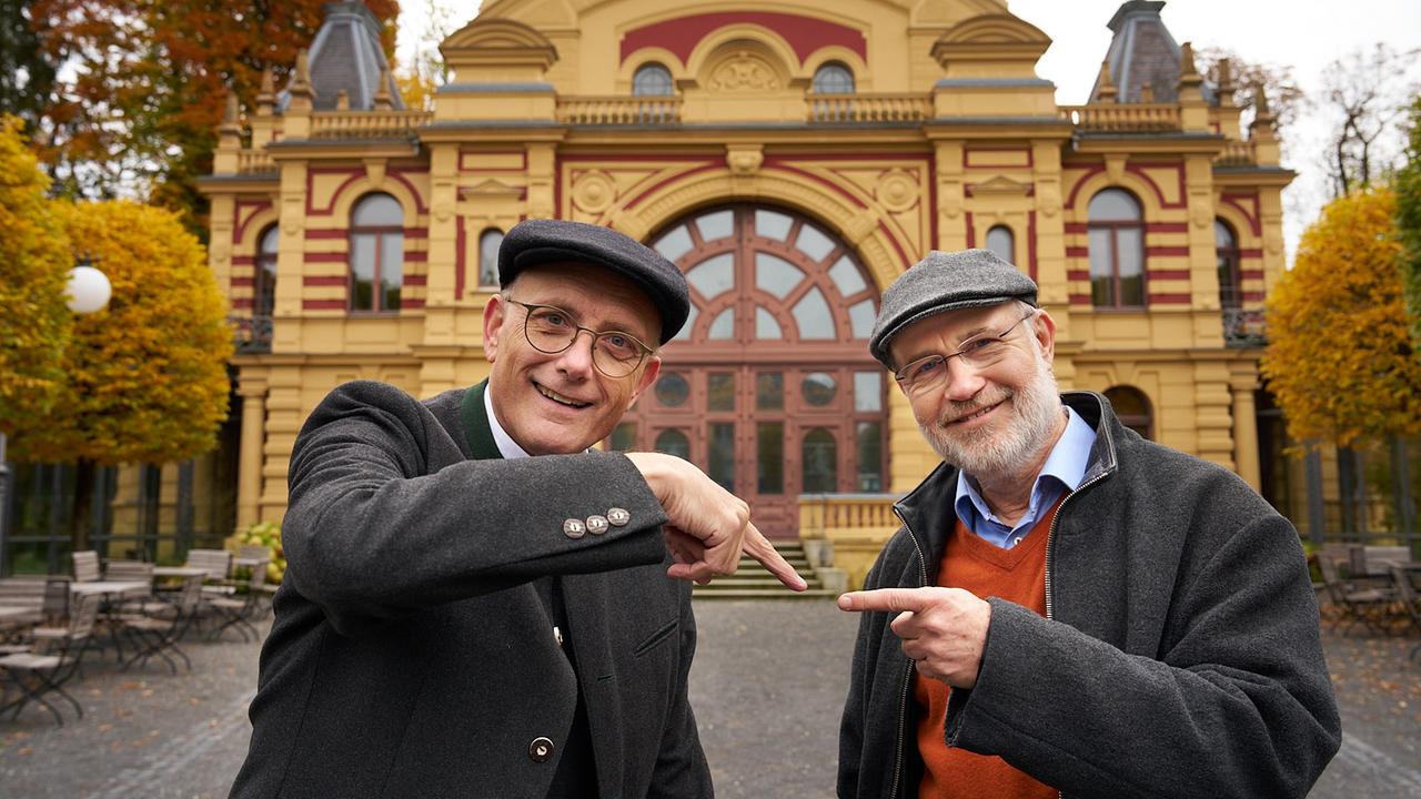 Thomas Schwartz und Harald Lesch stehen vor einem Theater. Beide schauen lächelnd in die Kamera. Jeder zeigt auf den jeweils anderen.