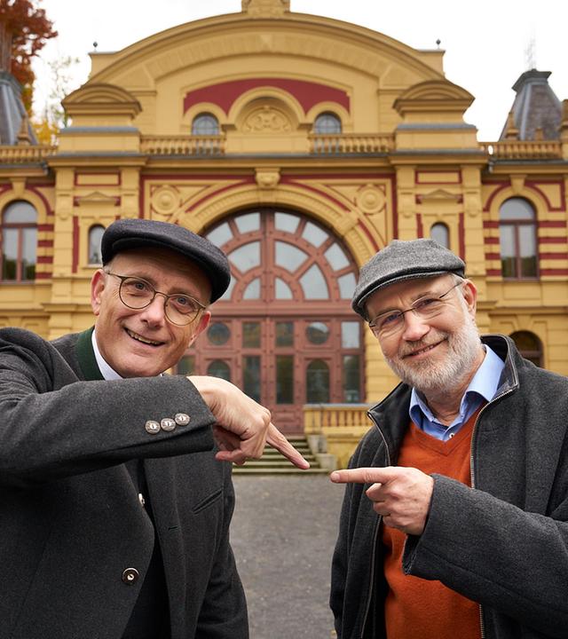 Thomas Schwartz und Harald Lesch stehen vor einem Theater. Beide schauen lächelnd in die Kamera. Jeder zeigt auf den jeweils anderen.